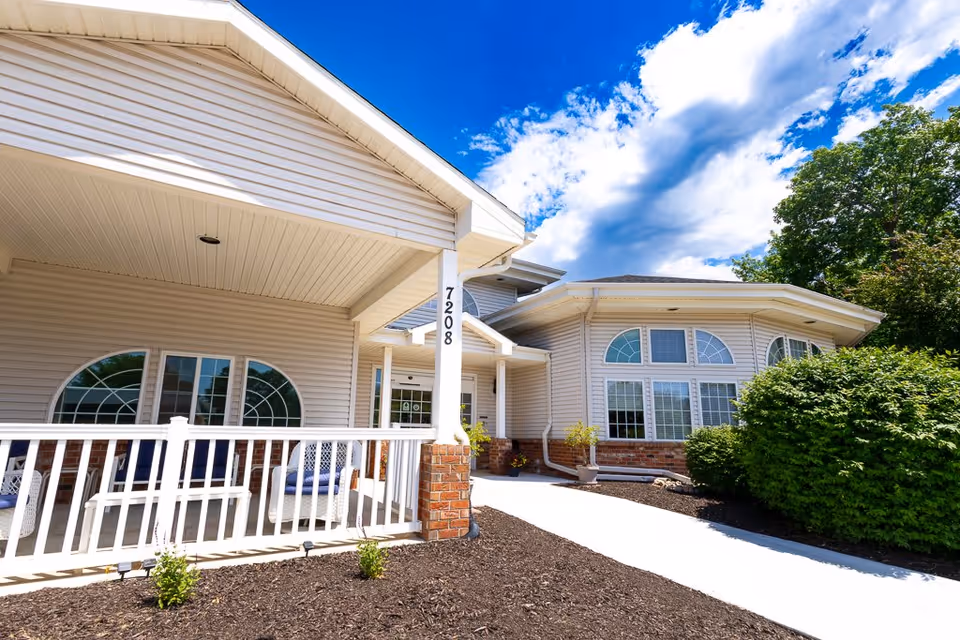 Exterior view of Bailey Pointe Assisted Living at Van Dorn showing the building entrance with white siding, large arched windows, a covered porch with white railing, and a clear blue sky with some clouds above.