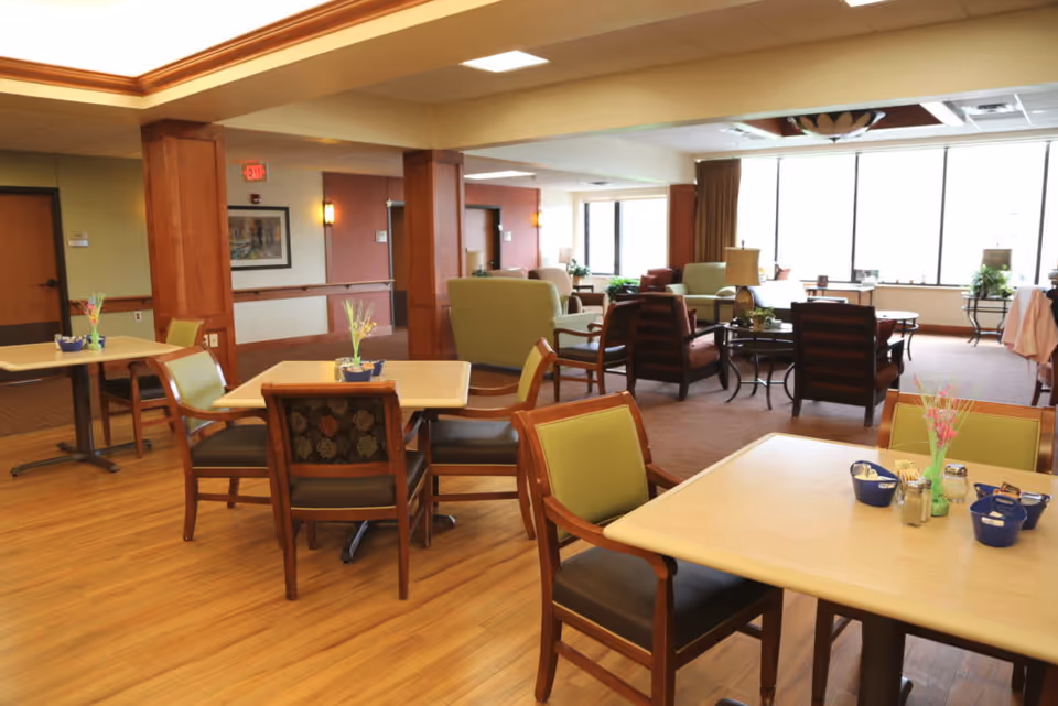 A spacious and well-lit common area in a senior living facility featuring several tables with chairs arranged for dining or socializing. The room has wooden flooring, large windows letting in natural light, and a seating area with armchairs and sofas in the background. Small decorative plants and condiment containers are placed on the tables.