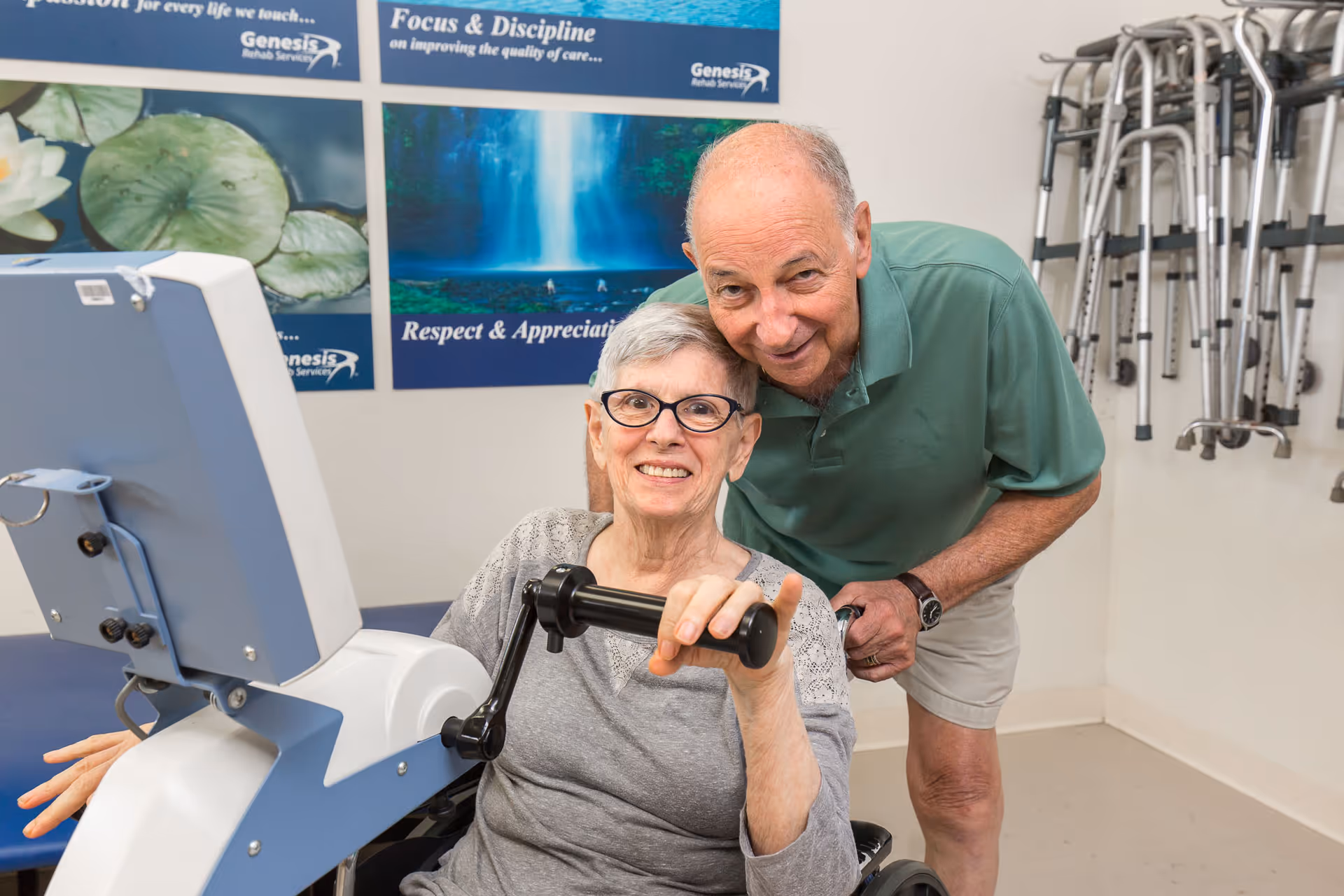 An elderly woman with glasses is seated and using a hand exercise machine while smiling. An elderly man in a green polo shirt and beige shorts is standing behind her, leaning forward and smiling. In the background, there are posters on the wall and a rack with multiple walkers.
