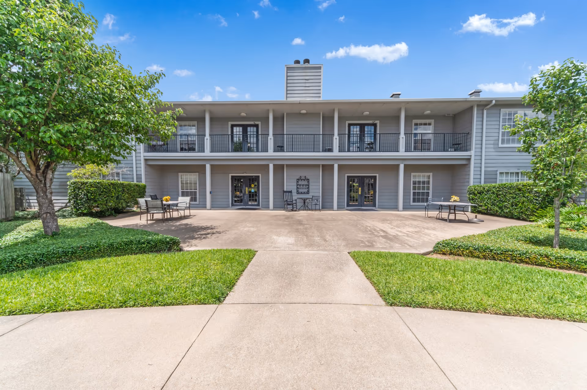 Exterior view of a two-story senior living facility building with gray siding, black railings, and multiple doors and windows. There is a concrete patio area with outdoor tables and chairs, surrounded by well-maintained green grass, bushes, and trees under a blue sky with scattered clouds.