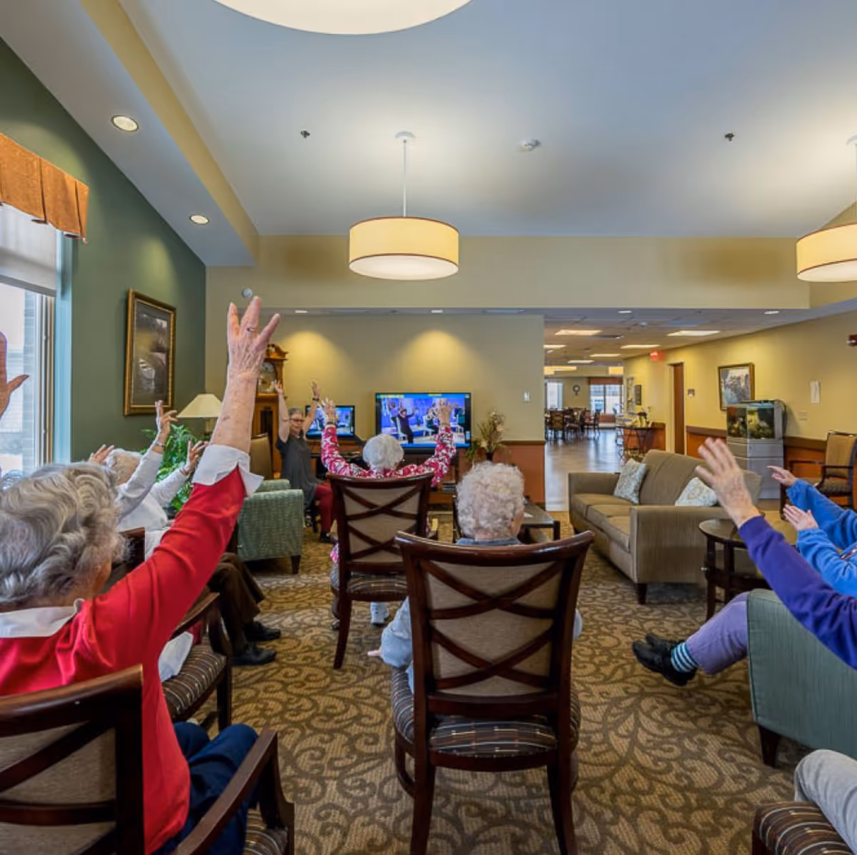 A group of elderly people seated in a living room area participating in a seated exercise or activity, raising their arms while facing a television screen. The room is well-lit with ceiling lights, has comfortable chairs, a sofa, and a patterned carpet. There are paintings on the walls and a fish tank in the background.