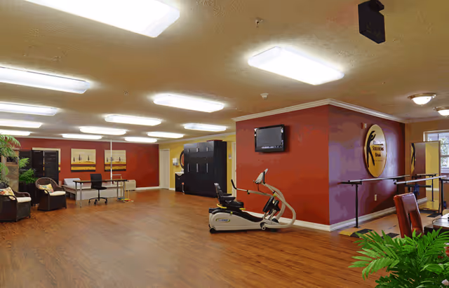 A spacious senior living facility room with wooden flooring and red walls. The room features exercise equipment including a recumbent bike and parallel bars for physical therapy. There are also chairs, a desk with a chair, and decorative paintings on the walls. A television is mounted on one wall, and there are plants adding greenery to the space.