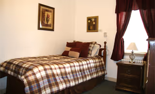 Bedroom in retirement center with plaid bed, nightstand, and dark drapes.