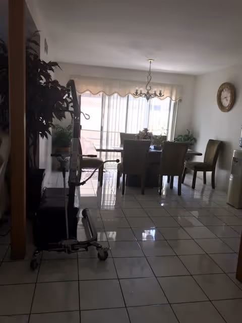 A dining room with a rectangular table surrounded by six chairs. There is a chandelier hanging above the table. Large sliding glass doors with vertical blinds allow natural light into the room. A clock is mounted on the wall to the right, and a water dispenser is visible nearby. The floor is tiled with white tiles, and there are some plants placed around the room.