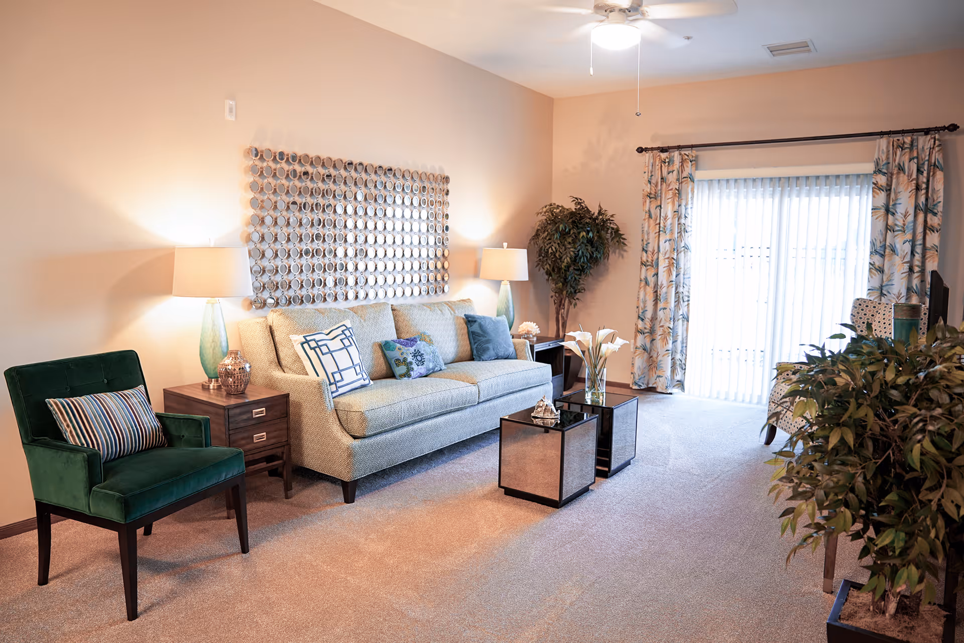 A cozy living room with a beige sofa adorned with decorative pillows, a green armchair with a striped pillow, two mirrored cube coffee tables with decorative items, two side tables with lamps, a large leafy plant in the corner, floral curtains covering a sliding glass door, and a ceiling fan overhead.