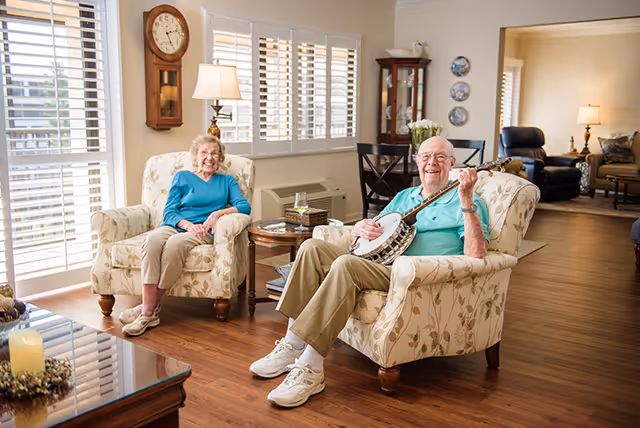 An elderly man and woman sitting in floral armchairs in a bright living room. The man is playing a banjo and smiling, while the woman is also smiling and sitting comfortably. The room has wooden floors, a wall clock, a lamp on a side table, and a glass-front cabinet with decorative items. In the background, there is another seating area with a lamp and armchairs.