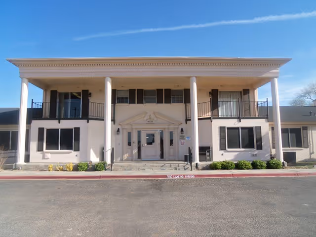 Two-story beige building with tall front columns, a central entrance and second-floor balconies under a clear blue sky.