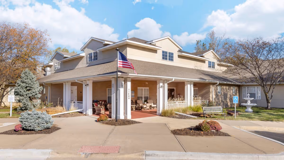 Front entrance of a senior living building with a covered porch, American flag, seating, fountain, and landscaped beds.