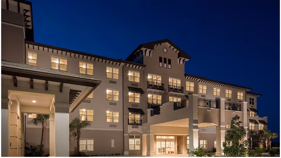 Exterior view of a multi-story senior living facility building at dusk with lights glowing from the windows and architectural details including balconies and columns.