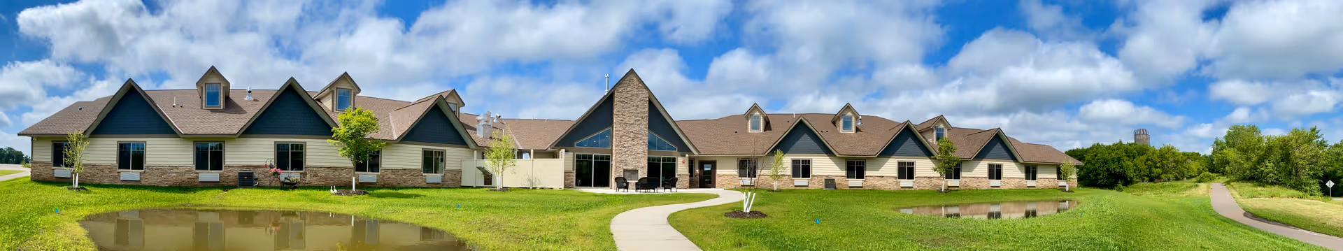 Panoramic front view of a single-story senior living building with gabled roofs, a central entrance, grassy lawns, walkways and small ponds under a partly cloudy sky.