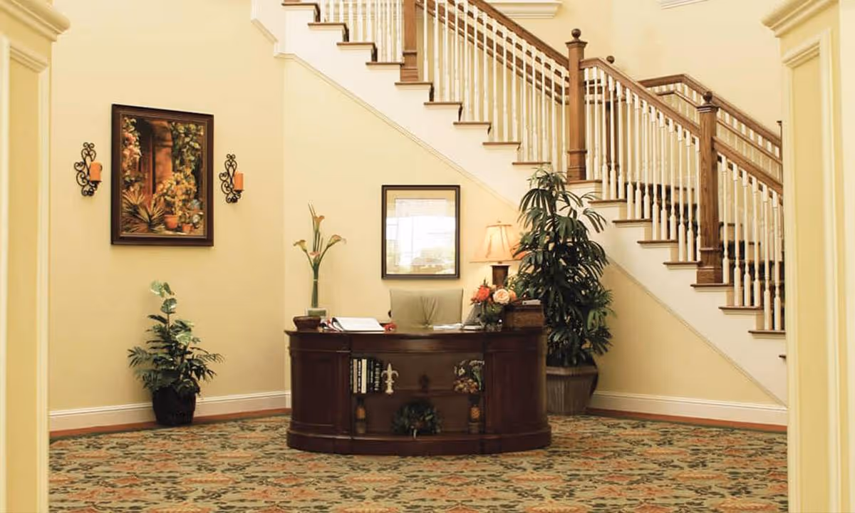 Reception area with a wooden desk placed in front of a staircase with white railings and wooden banisters. The desk has decorative items including flowers and books. The walls are painted light yellow, adorned with framed artwork and wall sconces with candles. There are potted plants on either side of the desk and a patterned carpet covering the floor.