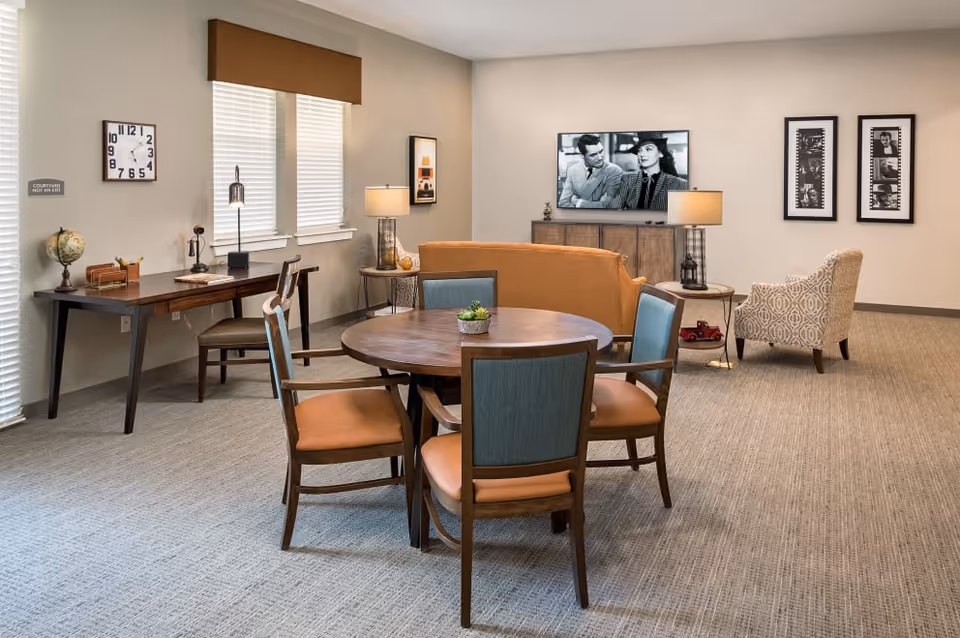 A cozy living room area with a round wooden table and four chairs in the foreground. Behind the table is a mustard-colored sofa facing a TV mounted on the wall displaying a black and white movie scene. To the right of the sofa is a patterned armchair and two framed black and white photos on the wall. On the left side of the room, there is a wooden desk with a chair, a globe, a clock on the wall, and two windows with blinds. The room has beige walls and carpeted flooring.