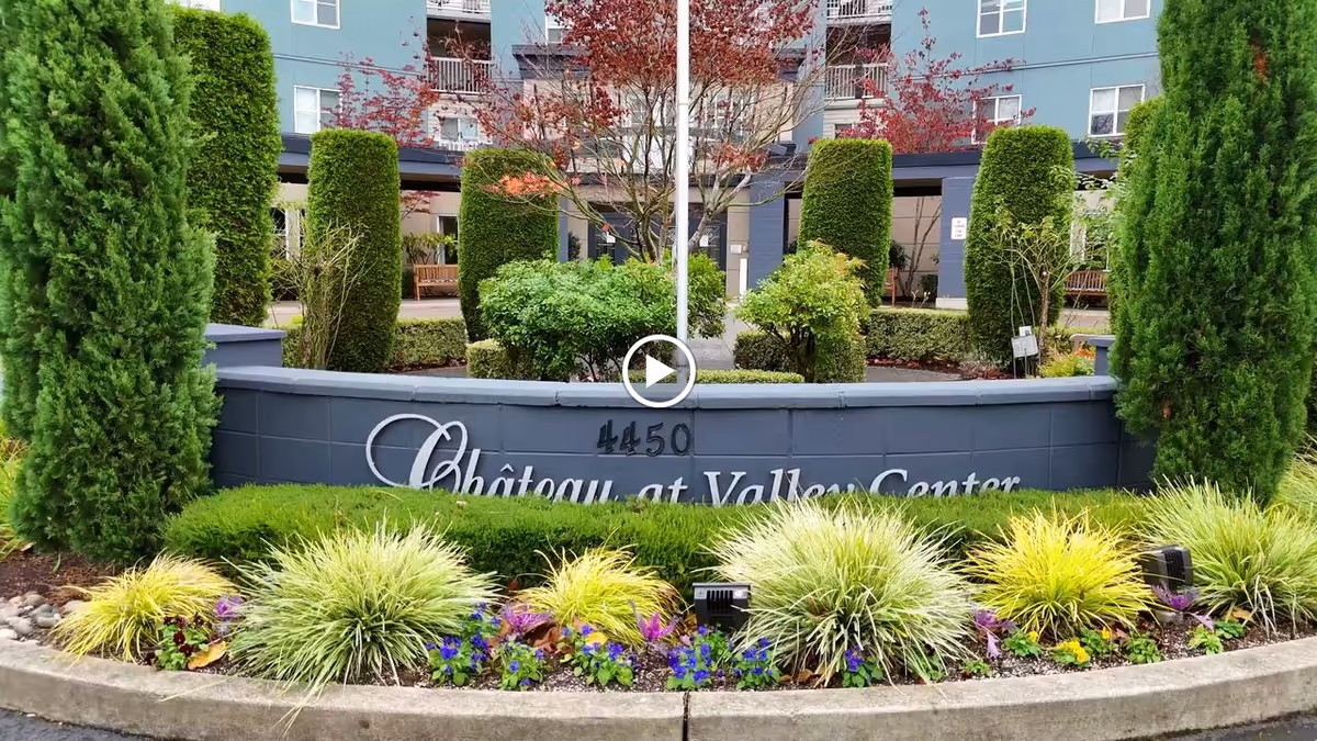 Entrance sign for Chateau at Valley Center surrounded by well-maintained landscaping including green shrubs, ornamental grasses, and small trees, with a multi-story building visible in the background.