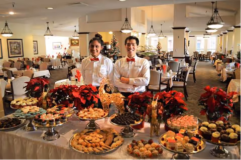 Two servers in white shirts and red bow ties stand behind a festive dessert buffet in a large dining room.