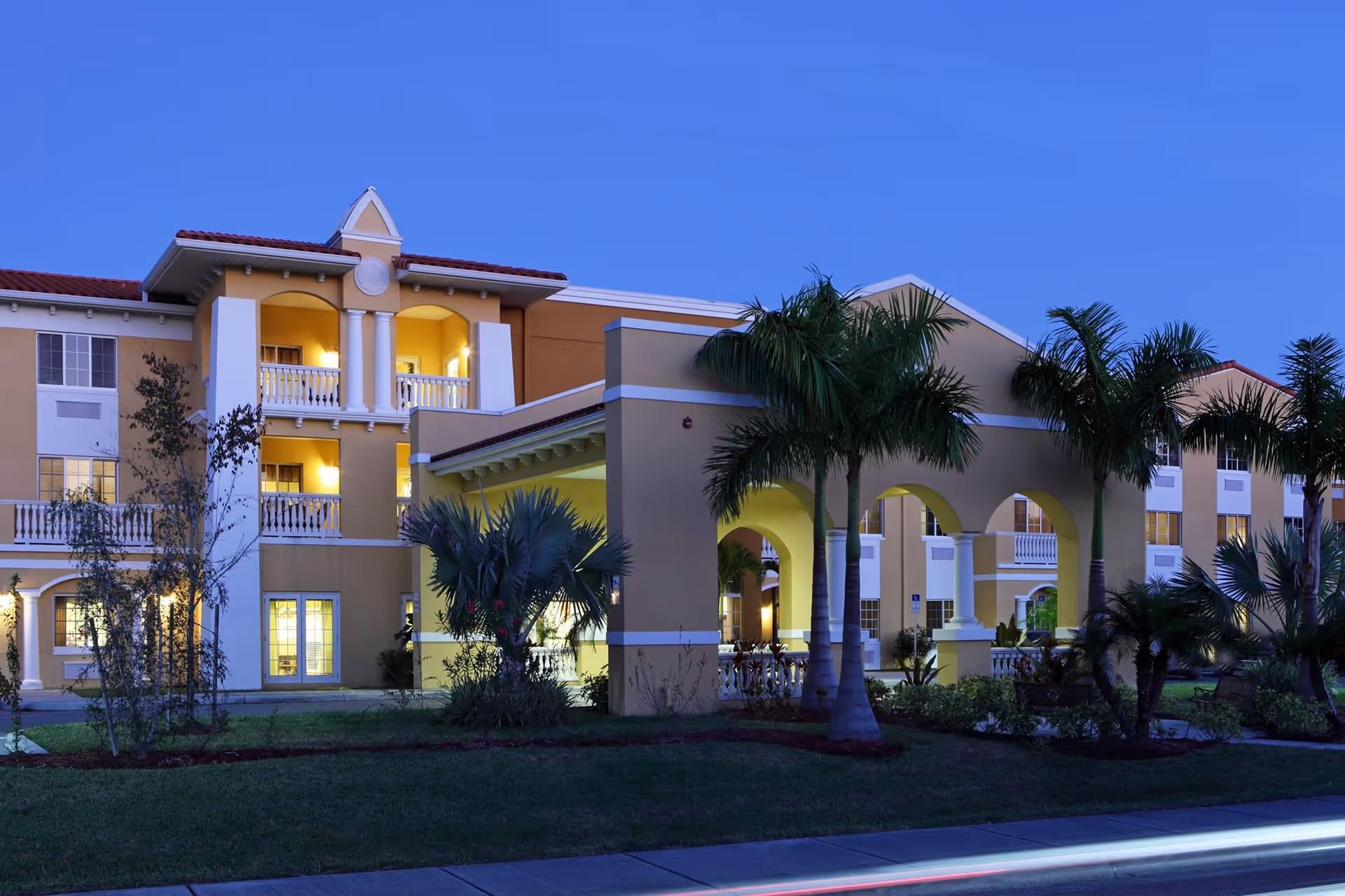 Exterior view of DeSoto Palms Assisted Living Community building at dusk, featuring a multi-story structure with balconies, arched entryways, palm trees, and well-maintained landscaping.