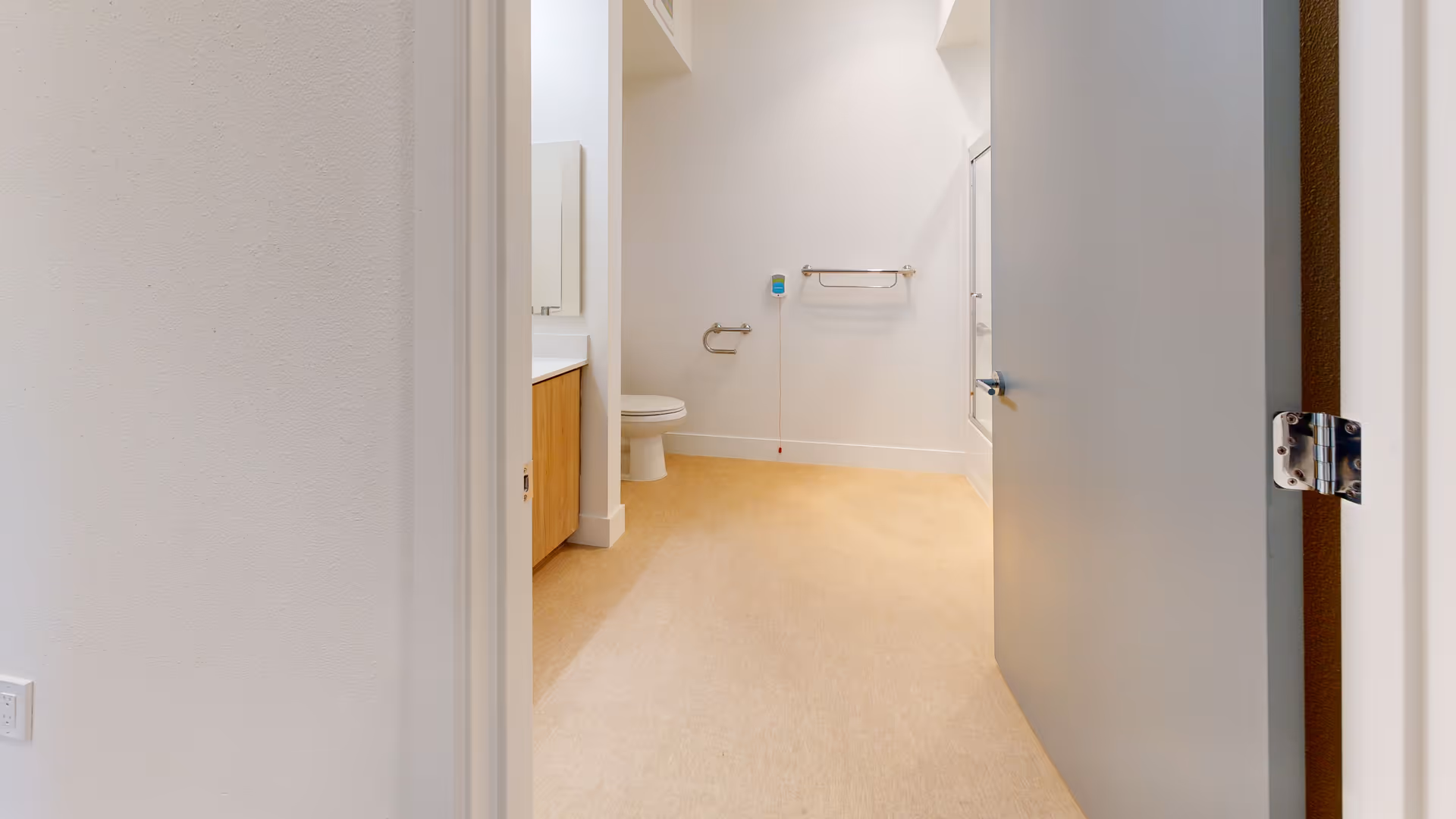 View into a clean, modern bathroom with beige flooring, white walls, a toilet, a wooden vanity with a white countertop, a mirror, a towel rack, and a bathtub with a glass door. The bathroom door is partially open.