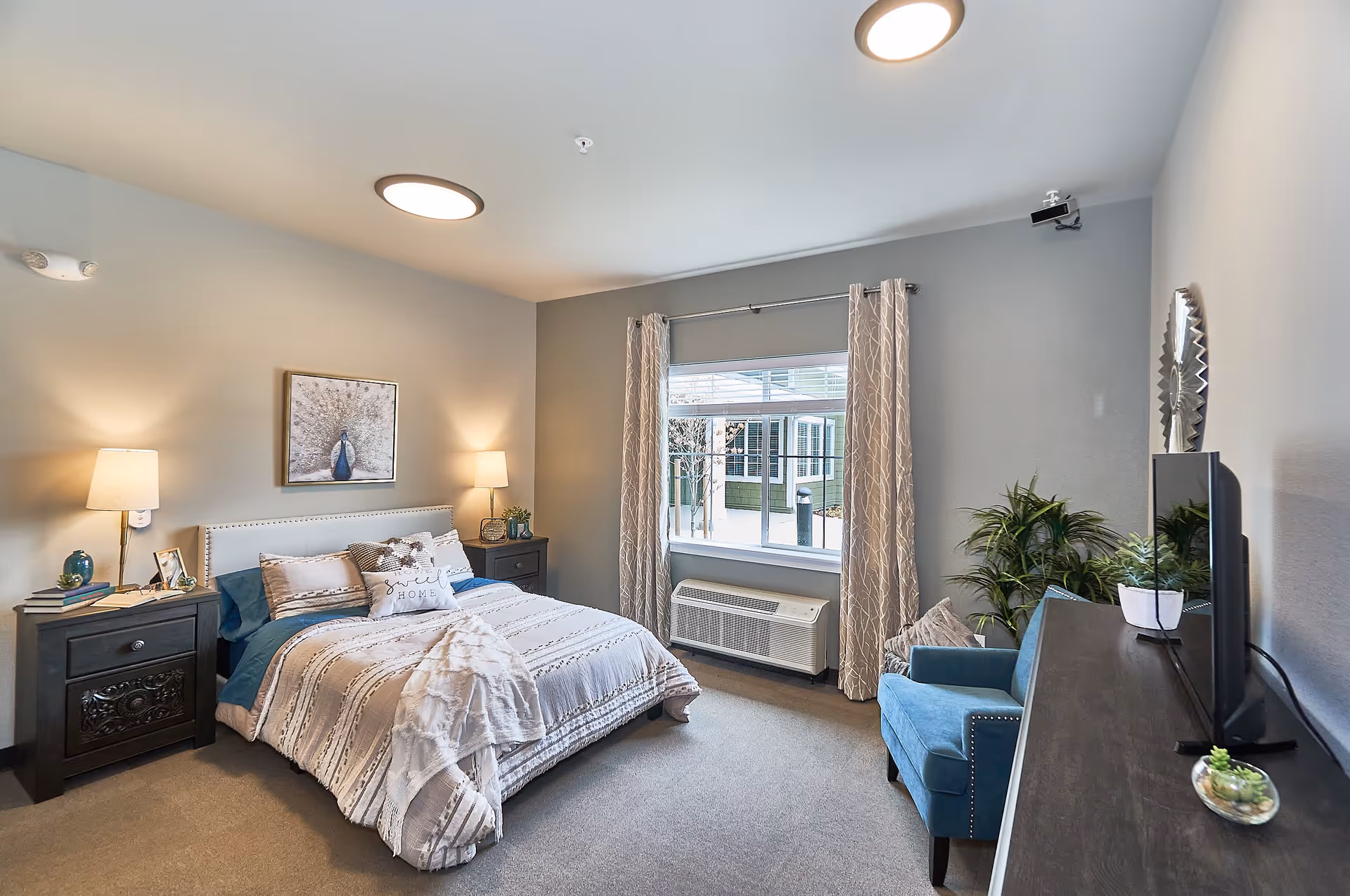 Bright, well-decorated bedroom with a made bed between two nightstands, a window with curtains, a blue armchair, and a TV on a dresser.