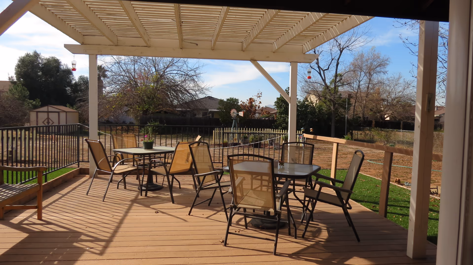 Outdoor patio area with a wooden deck covered by a pergola. There are two glass-top tables surrounded by several mesh chairs. A wooden bench is positioned on the left side. In the background, there is a fenced yard with trees, a small shed, and a windmill decoration under a partly cloudy sky.
