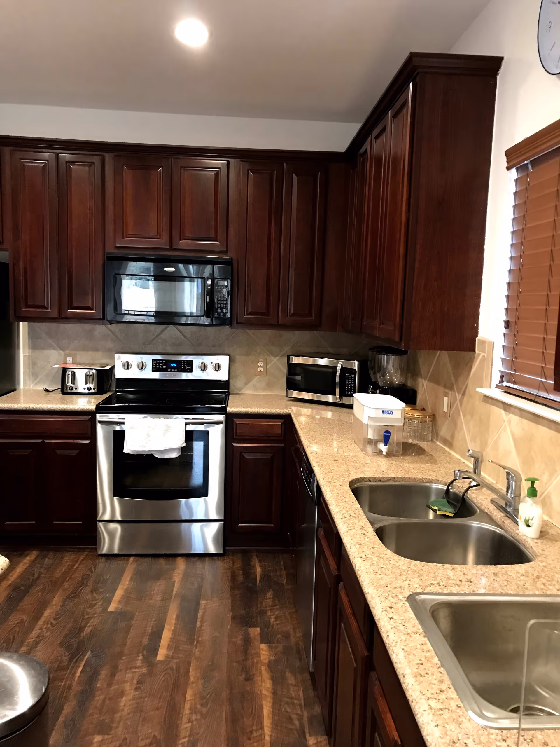 A kitchen with dark wooden cabinets, a stainless steel oven with a microwave above it, a toaster on the counter, a double sink, a dishwasher, a microwave, a blender, and a window with wooden blinds. The floor is wooden and the countertop is light-colored.
