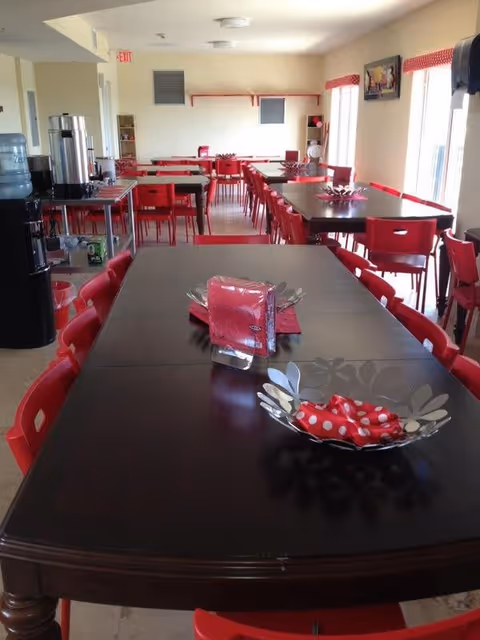 A dining room with multiple dark wooden tables and red chairs arranged neatly. Each table has a decorative metal bowl with red and white polka dot napkins and a napkin holder with red napkins. The room is well-lit with natural light coming through large windows on the right side. There is a coffee and water station on the left side near the entrance.