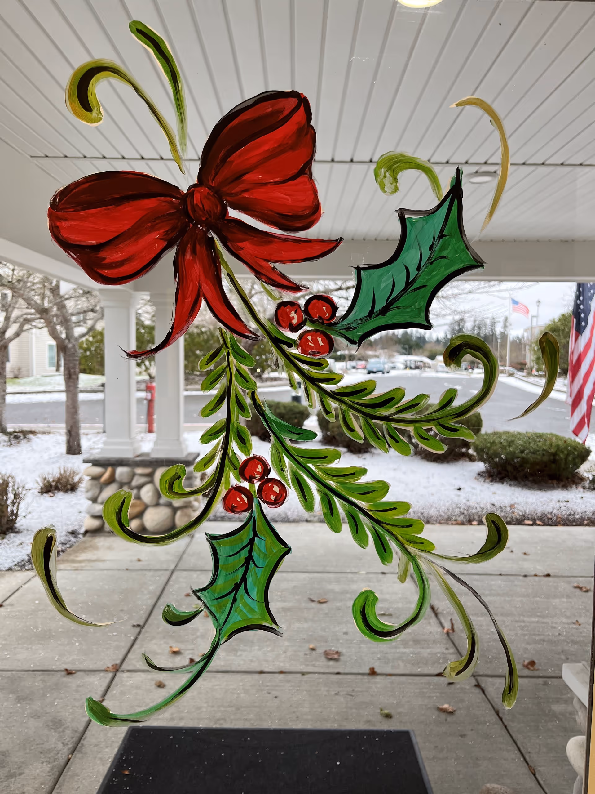 A festive holiday decoration painted on a glass window, featuring a large red bow, green holly leaves, red berries, and green pine branches. Outside the window, there is a covered walkway with white columns, a stone base, some bushes, trees, and a light dusting of snow on the ground. An American flag is visible in the background.