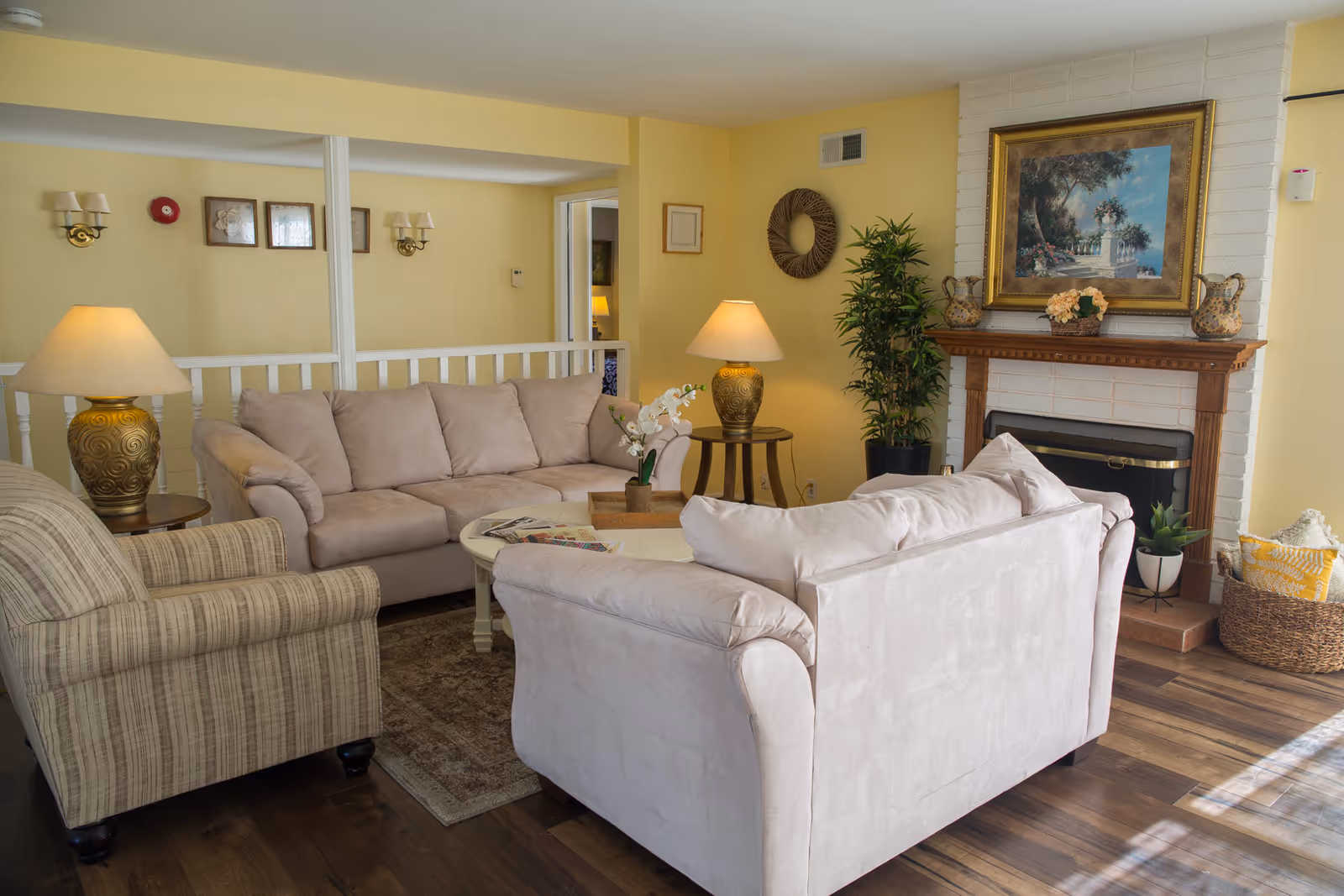 A cozy living room with two beige sofas and a striped armchair arranged around a white coffee table with magazines and a small orchid plant. The room has yellow walls, wooden flooring, two table lamps with ornate bases, a fireplace with a wooden mantel decorated with vases and a framed painting above it, and a basket with cushions near the fireplace.
