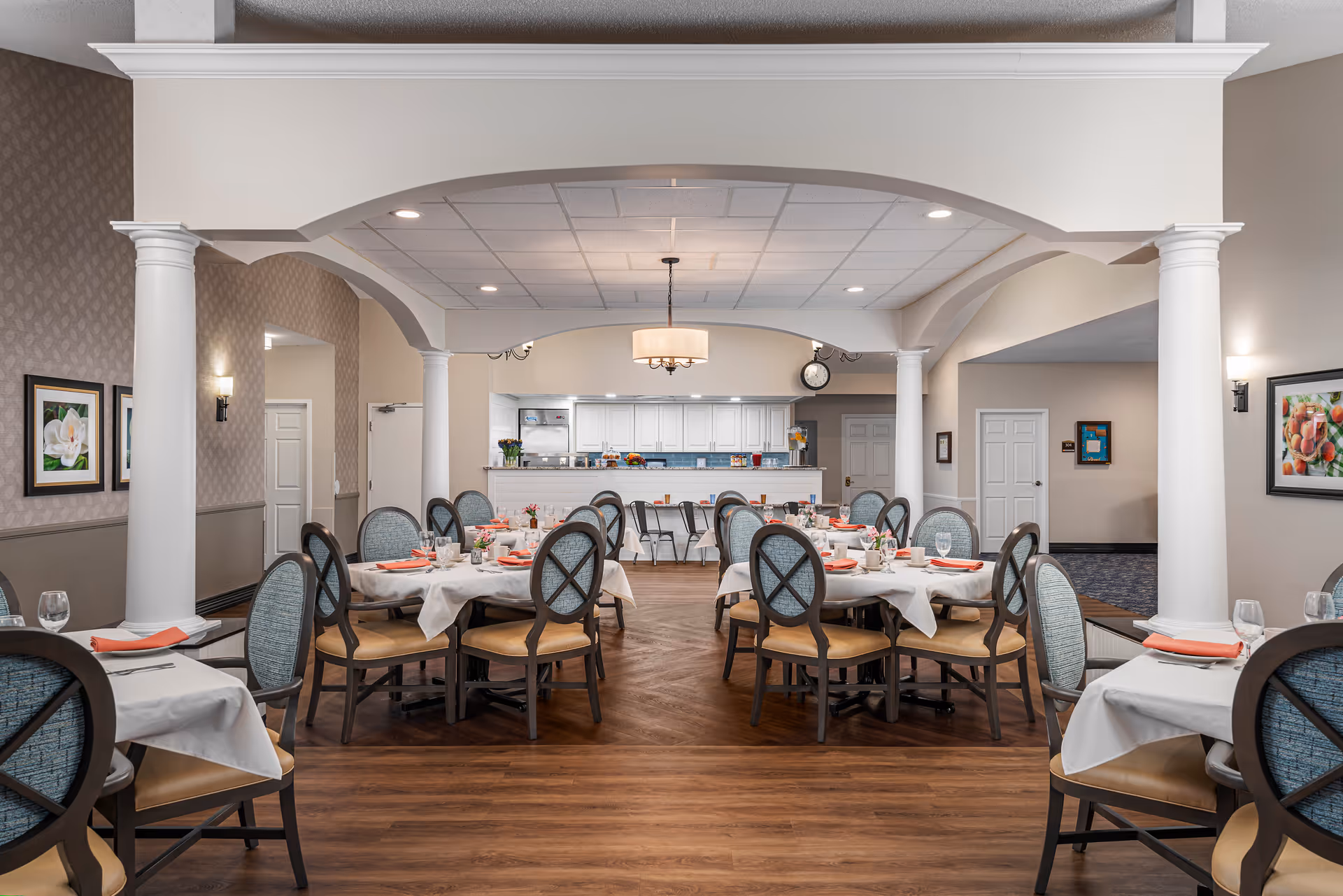 A well-lit dining room with multiple round tables covered with white tablecloths and set with plates, glasses, and orange napkins. The room features wooden flooring, white columns, and a ceiling with recessed lighting and a central hanging light fixture. The back of the room has a counter area with chairs and white cabinetry.