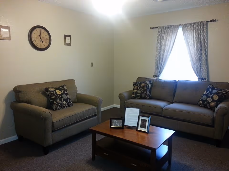 A cozy living room with a loveseat and a sofa, both upholstered in a neutral fabric with patterned throw pillows. A wooden coffee table with framed pictures and a document is placed in front of the seating. A window with patterned curtains allows natural light into the room. A wall clock and two small framed pictures hang on the beige wall.