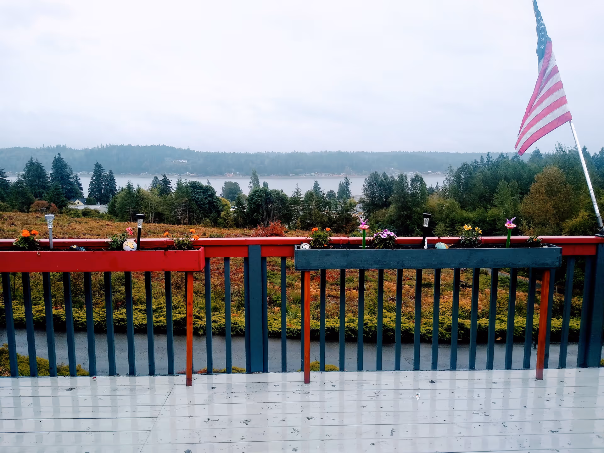 View from a painted wooden deck with railing and flower boxes overlooking trees and a body of water under a cloudy sky, an American flag at right.