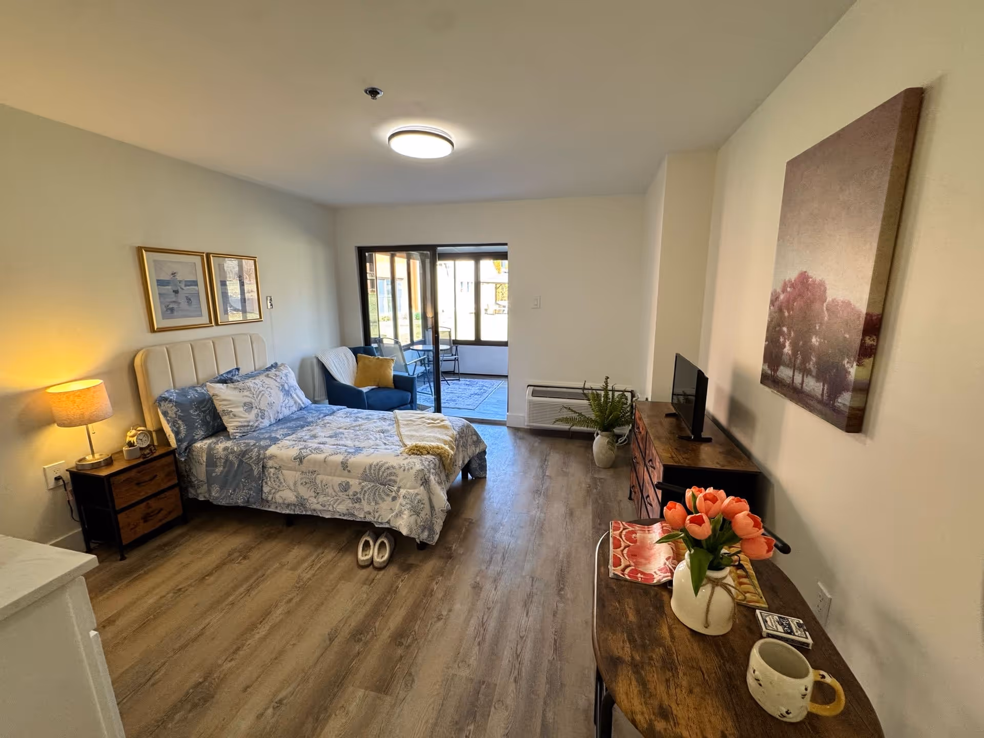 A cozy bedroom with a bed dressed in blue and white floral bedding, a nightstand with a lamp and clock, a blue armchair with a yellow pillow, a wooden dresser with a small TV, and a wooden table with a vase of pink tulips and a coffee mug. The room has wood flooring and a large window leading to a sunroom or enclosed patio area.