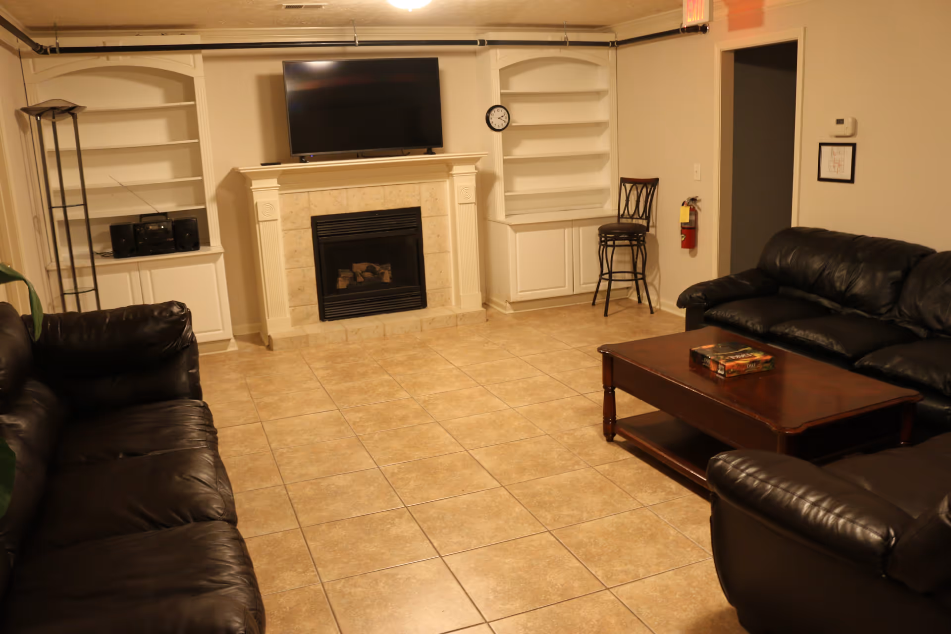 A cozy living room with two black leather sofas facing a wooden coffee table. A flat-screen TV is mounted above a fireplace with a tiled surround. There are built-in white shelves on either side of the fireplace, one with a stereo system. A tall floor lamp stands next to one shelf, and a single chair is placed near a doorway. The floor is covered with beige tiles.
