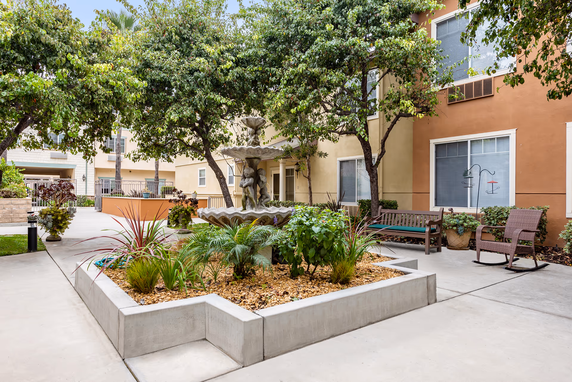 Outdoor courtyard area with a central stone fountain featuring cherub statues surrounded by plants and trees. There are benches and a rocking chair placed on the concrete pathway near the building with windows and beige walls.
