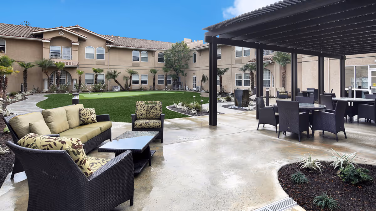 Outdoor courtyard area at Ivy Park at Simi Valley featuring patio furniture including cushioned chairs and sofas, a covered seating area with tables and chairs, a putting green, landscaped plants, and a two-story beige building with multiple windows in the background under a clear blue sky.