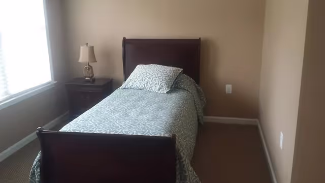 A small bedroom with a single bed covered in a patterned light blue and white bedspread and a matching pillow. Next to the bed is a dark wooden nightstand with a lamp. The room has beige walls and a window with white blinds letting in natural light.