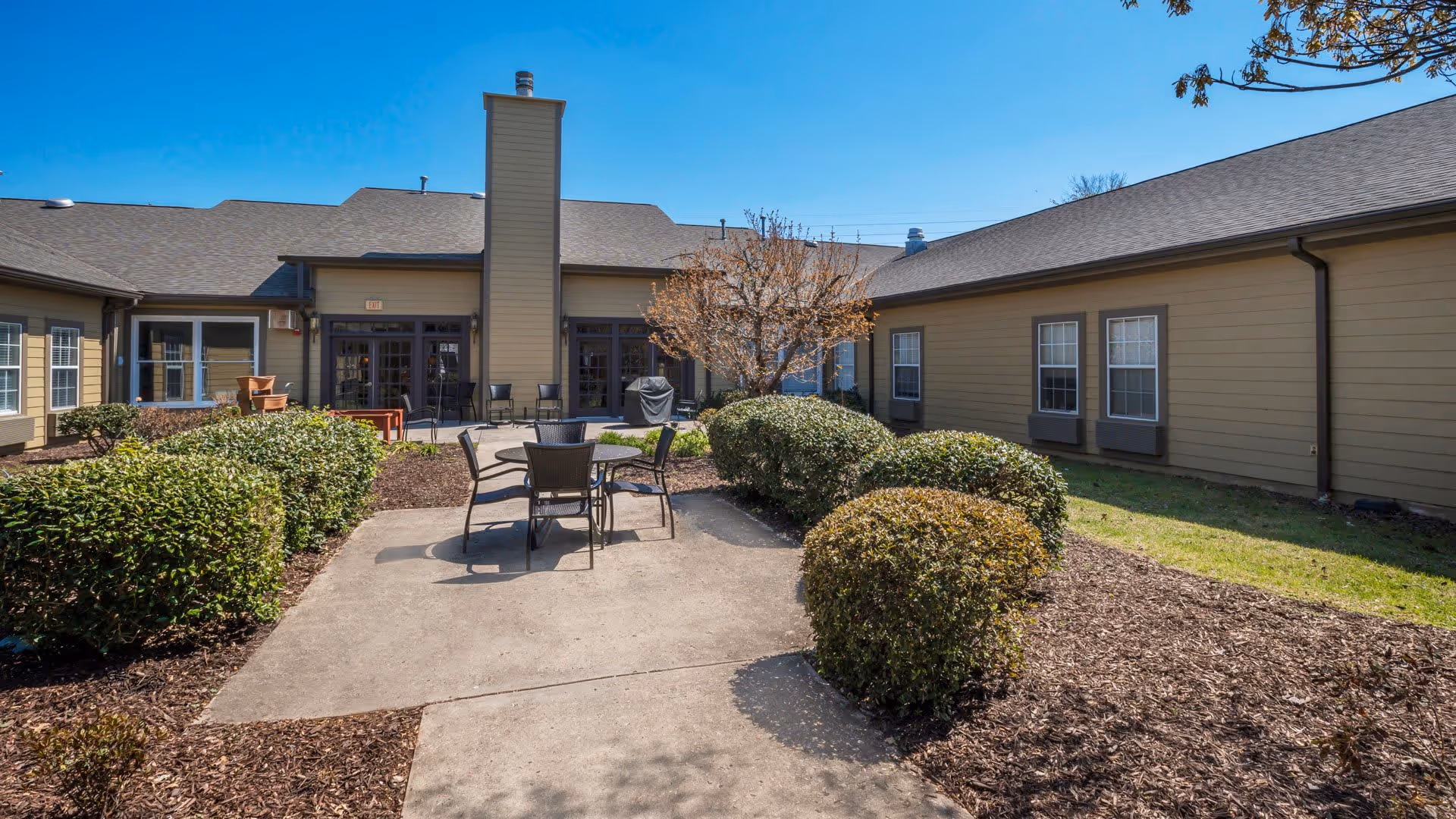 Outdoor courtyard area of a senior living facility with a concrete patio, several chairs and a table, surrounded by trimmed bushes and a small tree. The building exterior is beige with multiple windows and a central chimney. The sky is clear and blue.