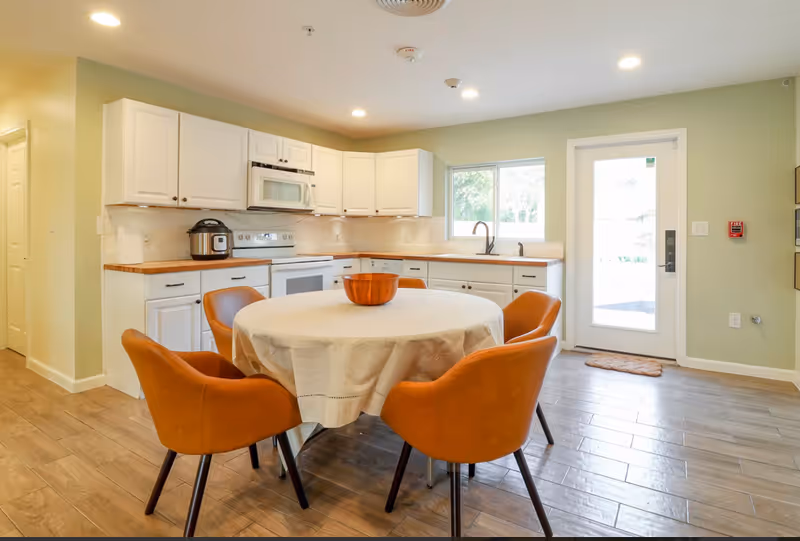 A bright kitchen with white cabinets, a microwave above the stove, and a round dining table covered with a beige tablecloth surrounded by four brown chairs. There is a window above the sink and a glass door leading outside, with wooden flooring throughout.