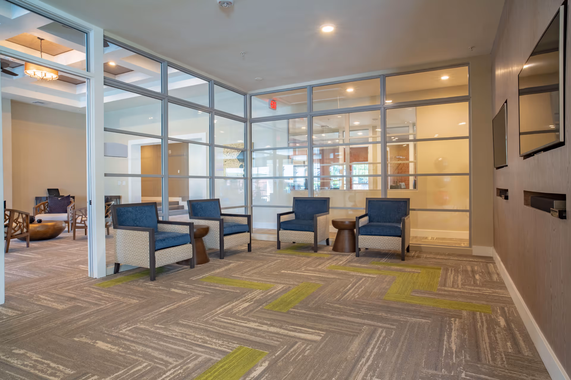 A modern lounge area with four blue cushioned armchairs arranged around two small wooden side tables. The room features large glass windows and doors with metal frames, allowing a view into an adjacent room with additional seating. The floor is carpeted with a patterned design in shades of gray and green. Two flat-screen televisions are mounted on the wall to the right.