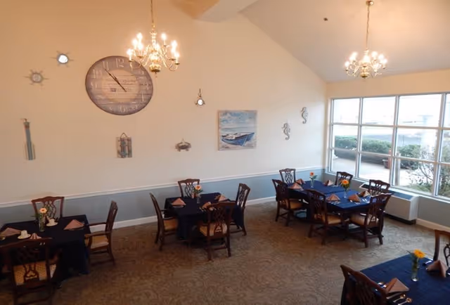 Dining room with several tables covered in dark blue tablecloths, each set with folded napkins and small flower vases. The room has high ceilings with two chandeliers, large windows letting in natural light, and nautical-themed wall decorations including a large clock, oars, and paintings of boats.