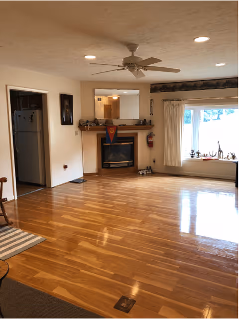 A spacious living room with polished wooden floors, a ceiling fan, and a fireplace with a mirror above it. To the left, there is an open doorway leading to a kitchen area with a refrigerator visible. On the right side, there is a large window with white curtains and decorative items on the windowsill. A fire extinguisher is mounted on the wall near the fireplace.