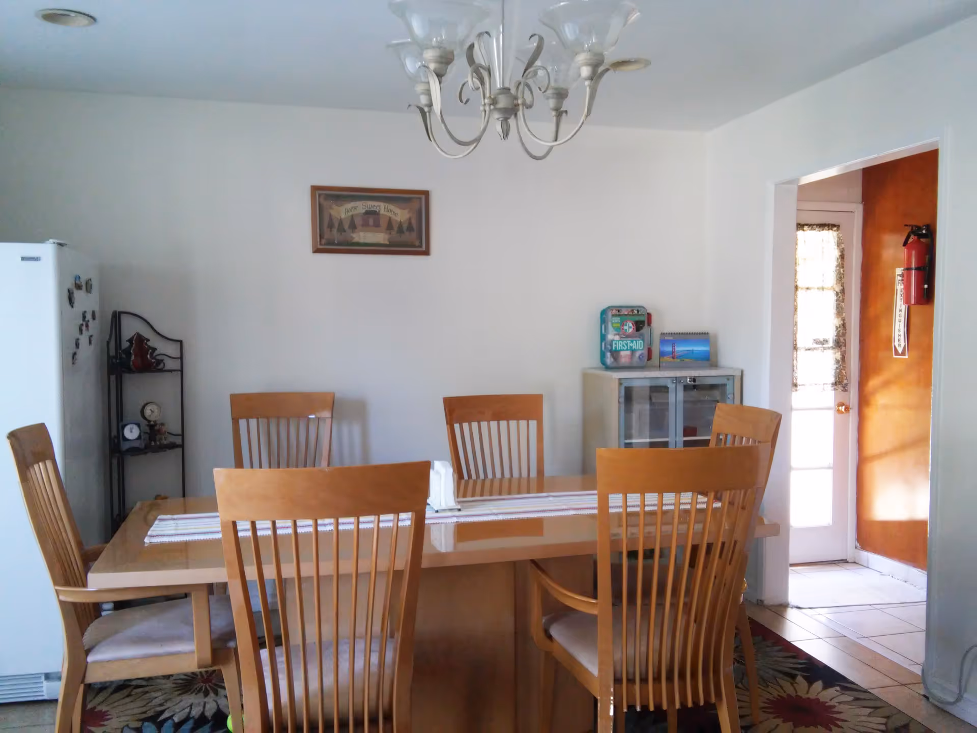 Dining room with a wooden table and six chairs, chandelier overhead, a refrigerator on the left and a doorway to the right.