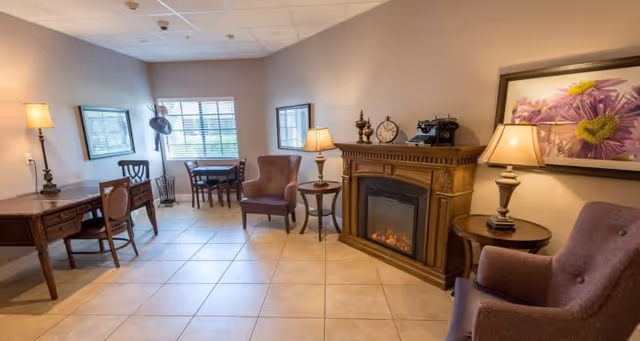 A cozy common room with upholstered armchairs, wooden desks and tables, lamps, and an electric fireplace beneath framed artwork.