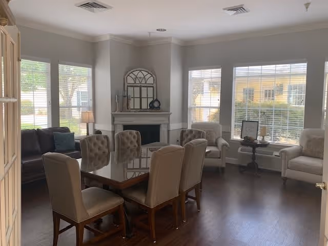 A well-lit living room with large windows featuring white blinds. The room contains a dark wooden dining table with six beige upholstered chairs around it. There is a fireplace with decorative items on the mantel, including a mirror and a clock. To the right, there are two beige armchairs with a small round table between them, holding a framed sign and a candle. A dark brown sofa with a green cushion is visible on the left side near the windows.
