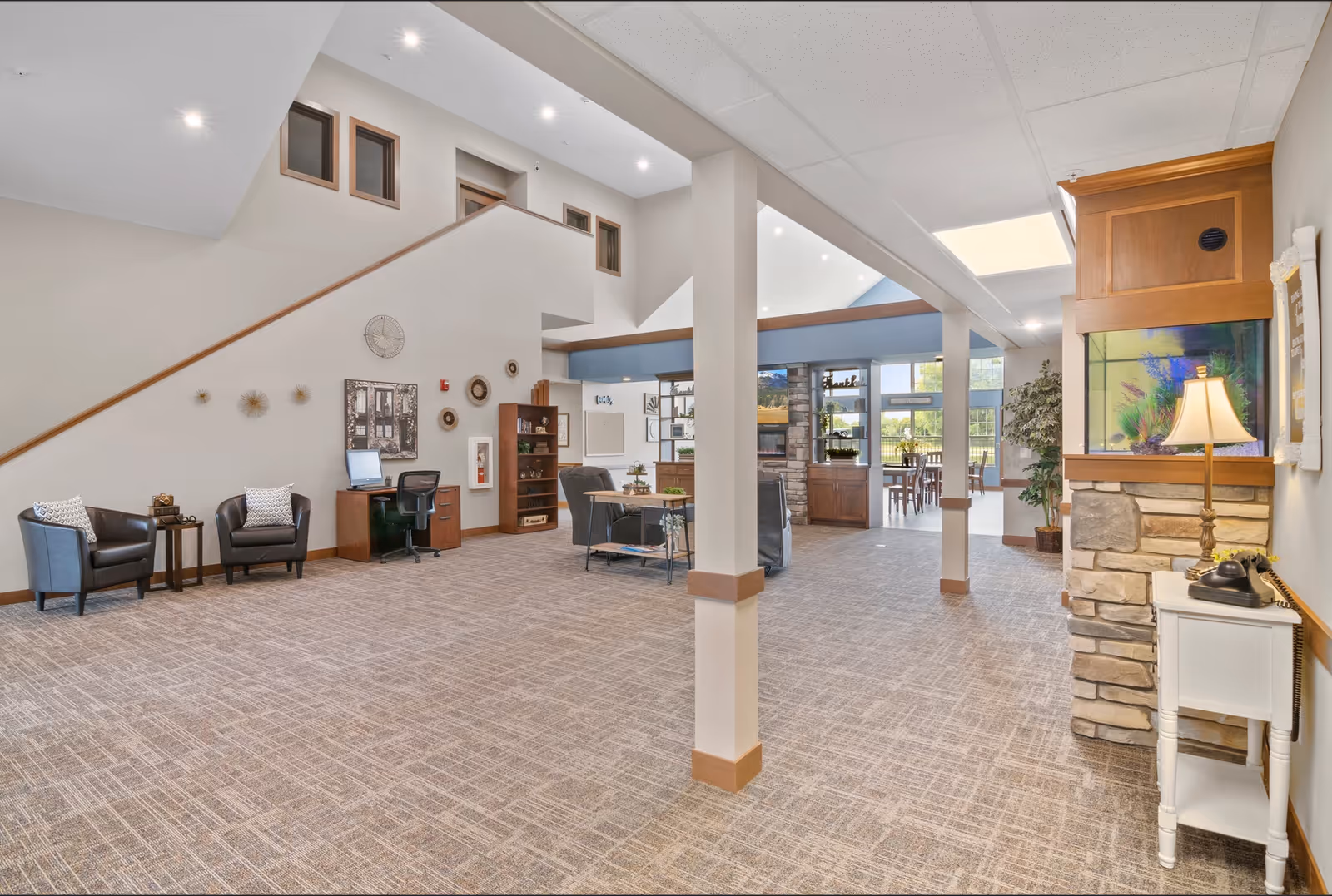 Spacious senior living common area with seating, columns, a staircase, and an open dining area visible in the background.