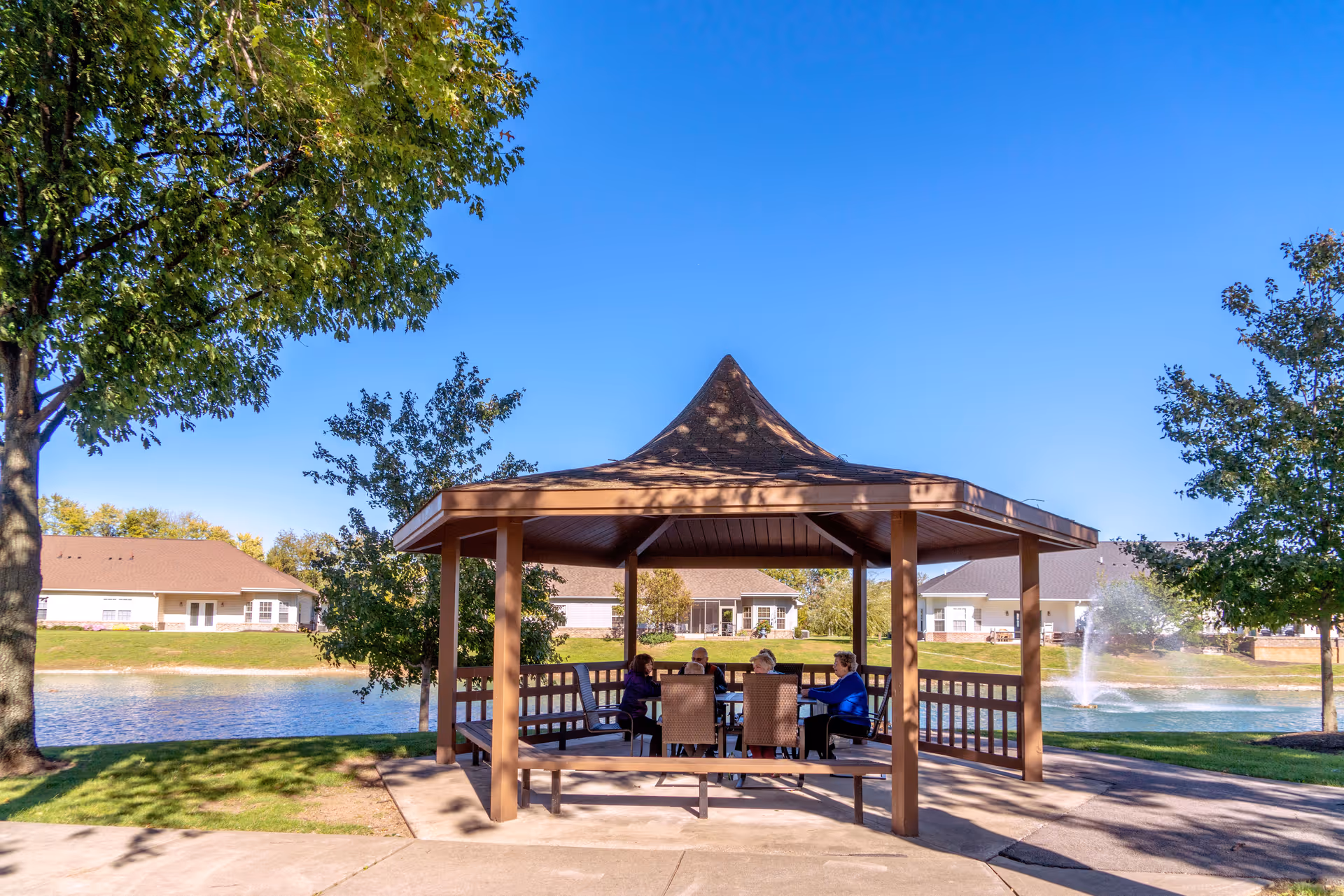 Several people seated under a wooden gazebo beside a pond with a fountain and nearby buildings.