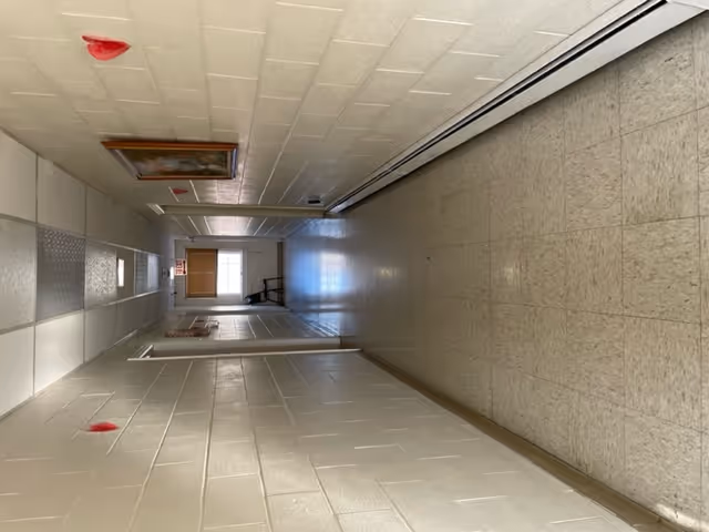 A long, empty hallway with beige tiled floor and white painted brick walls. The ceiling has white tiles with fluorescent lights and red fire alarm devices. There is a framed picture on the left wall and a window with blinds at the far end of the hallway.