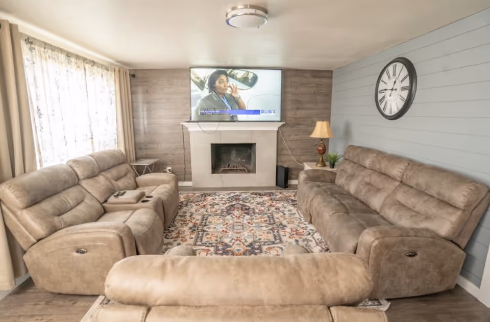A cozy living room with three beige recliner sofas arranged around a patterned area rug. A flat-screen TV is mounted above a fireplace on a wooden accent wall. There is a large clock on the light blue shiplap wall and a table lamp on a side table next to one of the sofas. Natural light comes in through a window with sheer curtains.