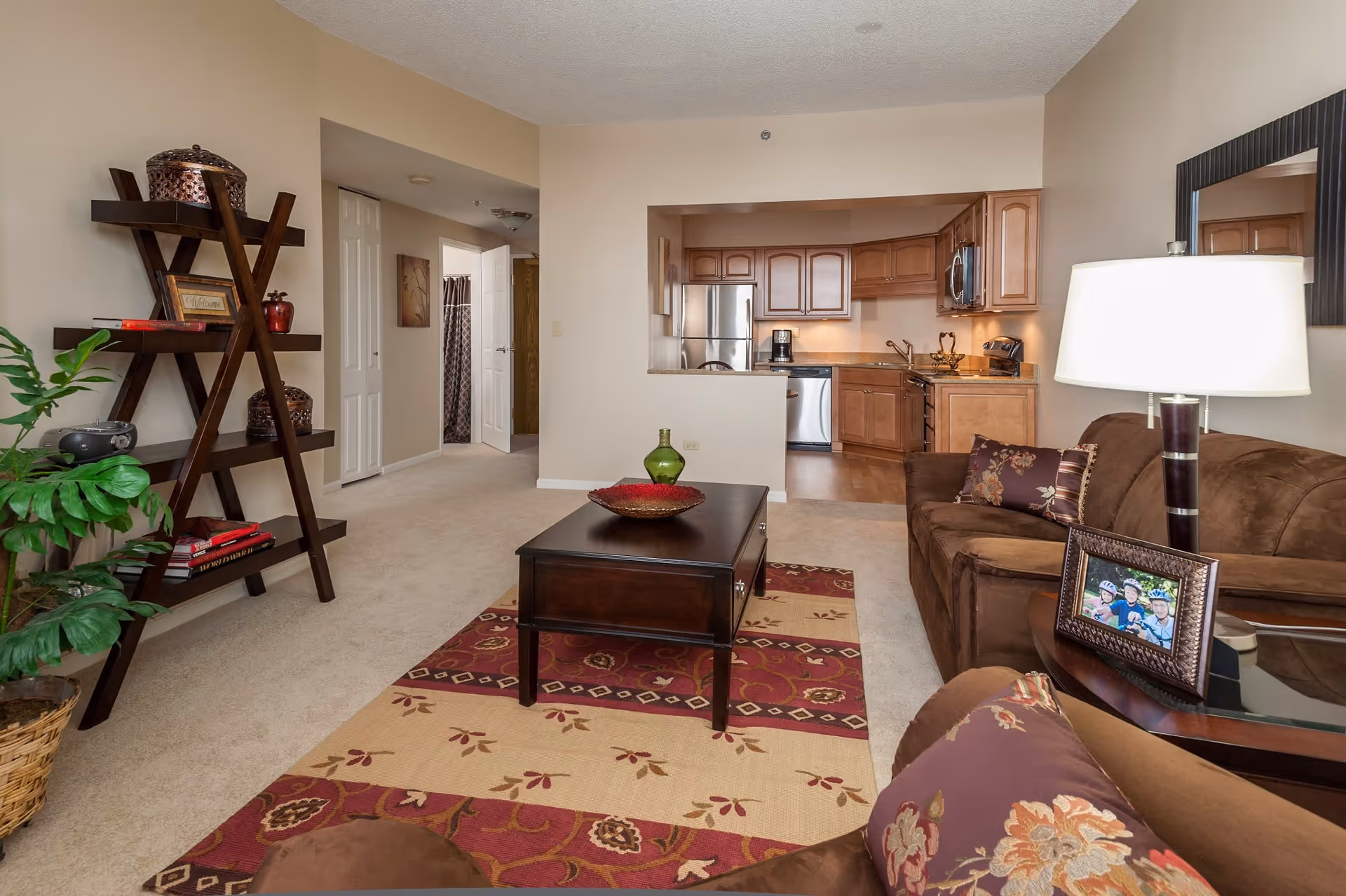 Open-plan living room with brown sofas, a coffee table on a patterned rug, decorative shelving and a view into a kitchen with wooden cabinets.