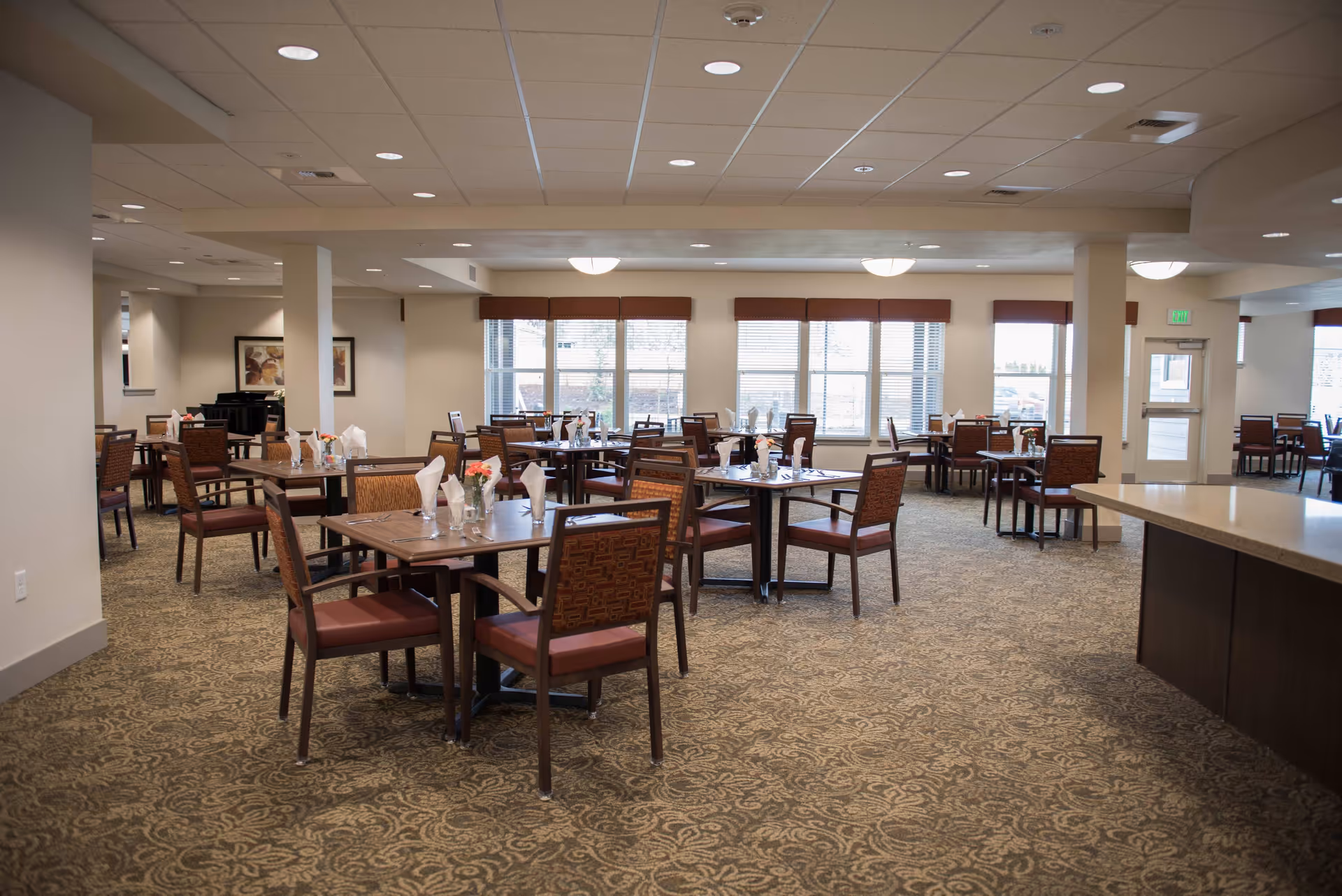 Well-lit dining room with multiple wooden tables and chairs set with napkins and flowers, large windows and patterned carpet.