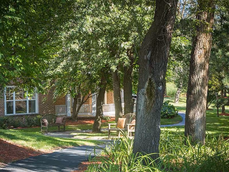 A peaceful outdoor garden area at Atria Park of Glen Ellyn with a paved walking path, several trees providing shade, green grass, and outdoor seating including chairs and benches near a brick building with windows and a porch.