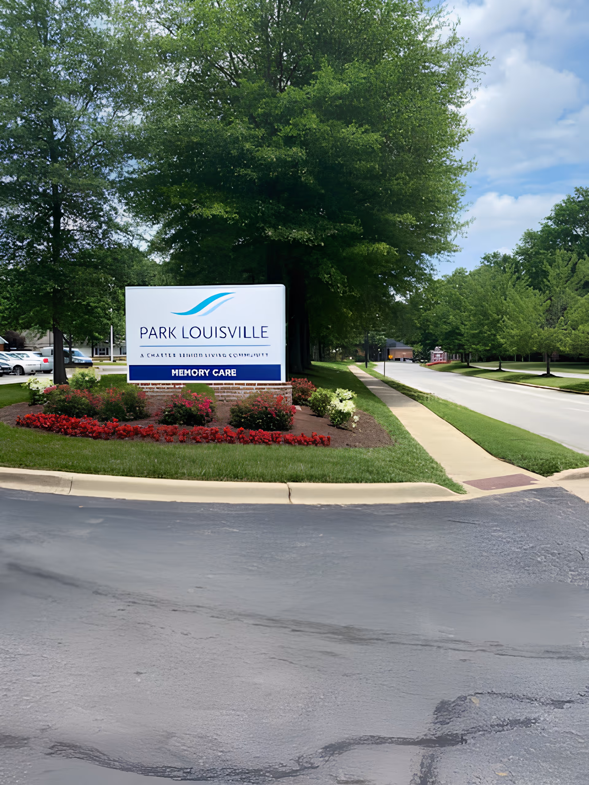 Outdoor view of a landscaped entrance area with a sign that reads 'Park Louisville, A Charter Senior Living Community, Memory Care' surrounded by green trees, grass, and flower beds under a partly cloudy sky.